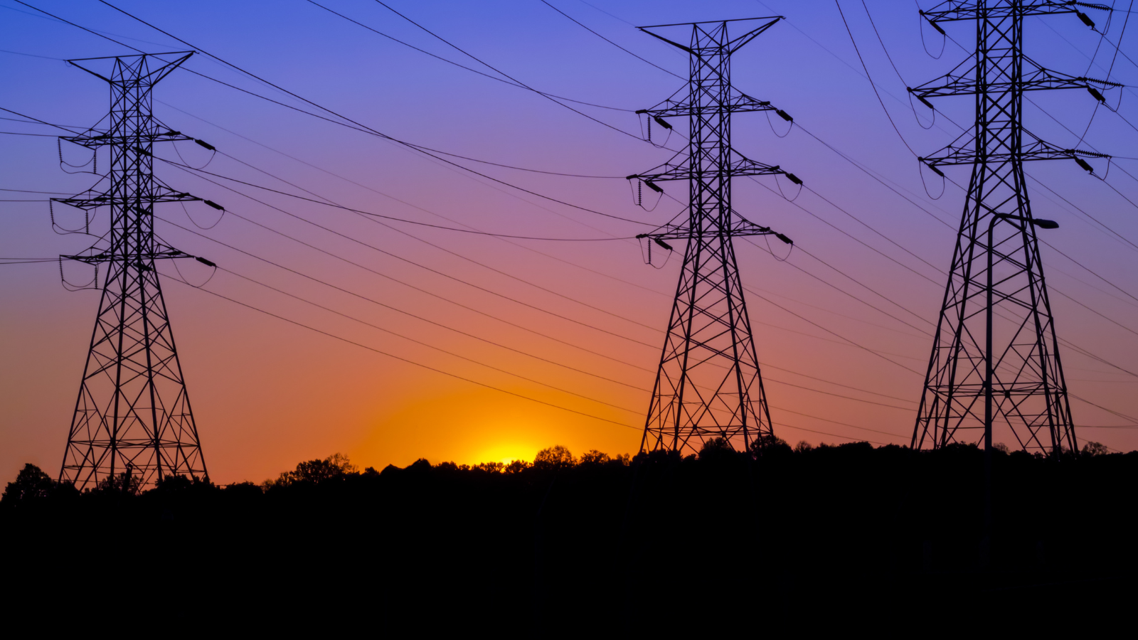 Stock image of transmission towers with the sun setting behind them. The sky is a purple, pink, and orange gradient.