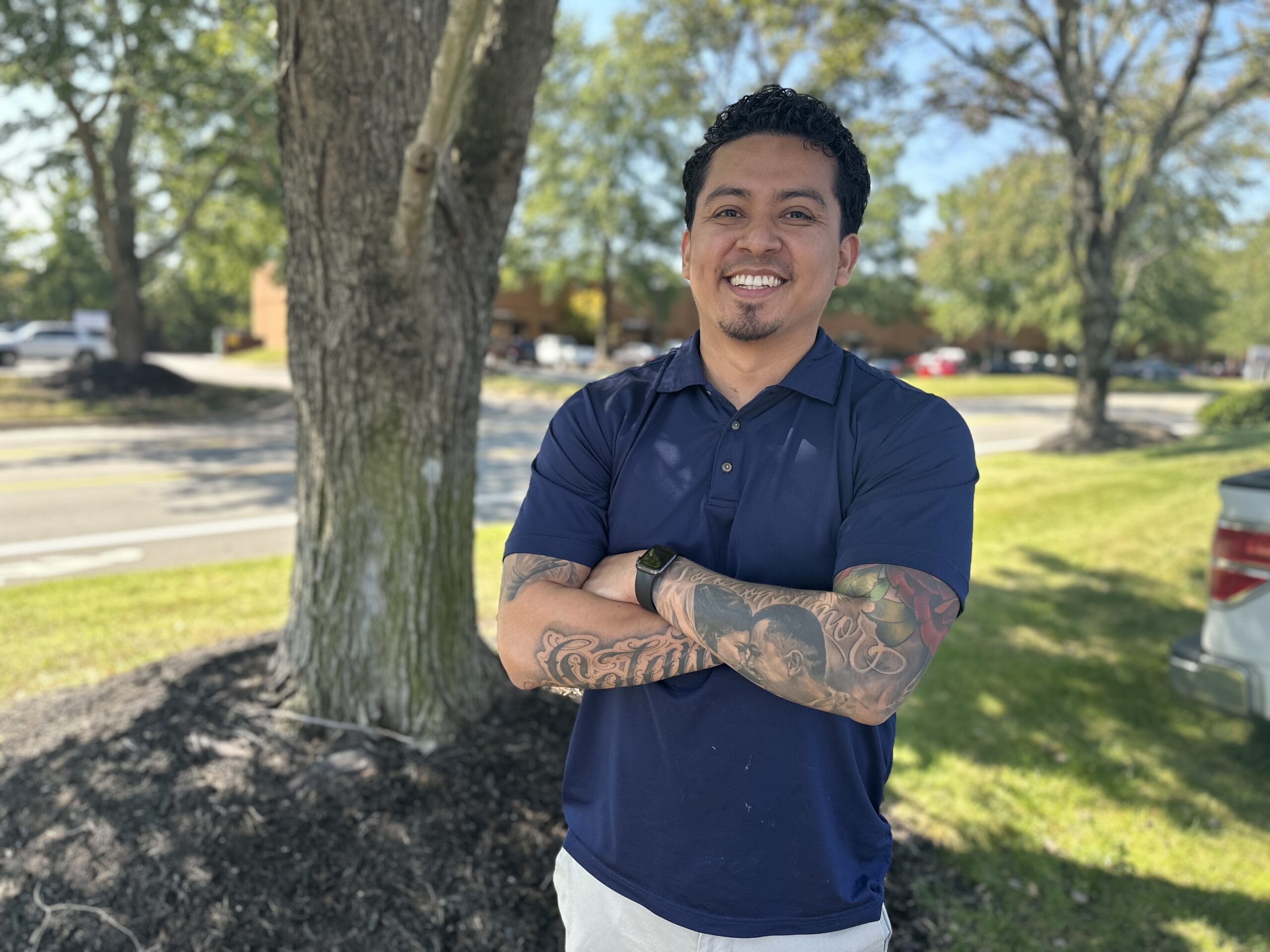 Carlos is dressed in a navy blue polo and light khaki pants and standing proudly against the backdrop of a tall tree with his arms crossed displaying his intricate tattoos.
