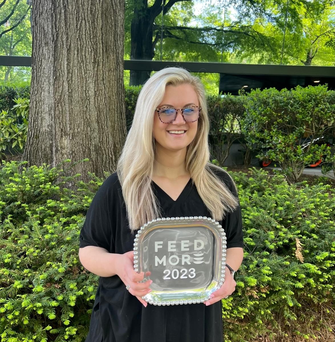 Photo of Taylor Dalton standing in front of a tree and shrubs wearing a black shirt and holding the Feed More 2023 award.