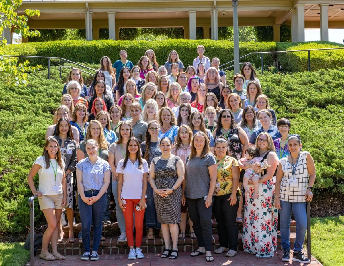 70 plus women taking a group photo standing on a brick staircase outside on a sunny day surrounded by green vegetation.