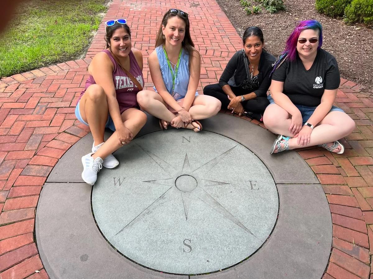 Alicia and three other women sitting on a red brick walkway posing with a gray stone compass embedded in the walkway.