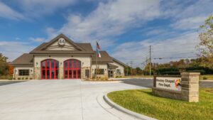 A building with the sign "Wesley Chapel Fire Dept." on the front, featuring a flag. Surrounding elements include trees, grass, and an overcast sky. The setting suggests an outdoor environment during autumn. Copy