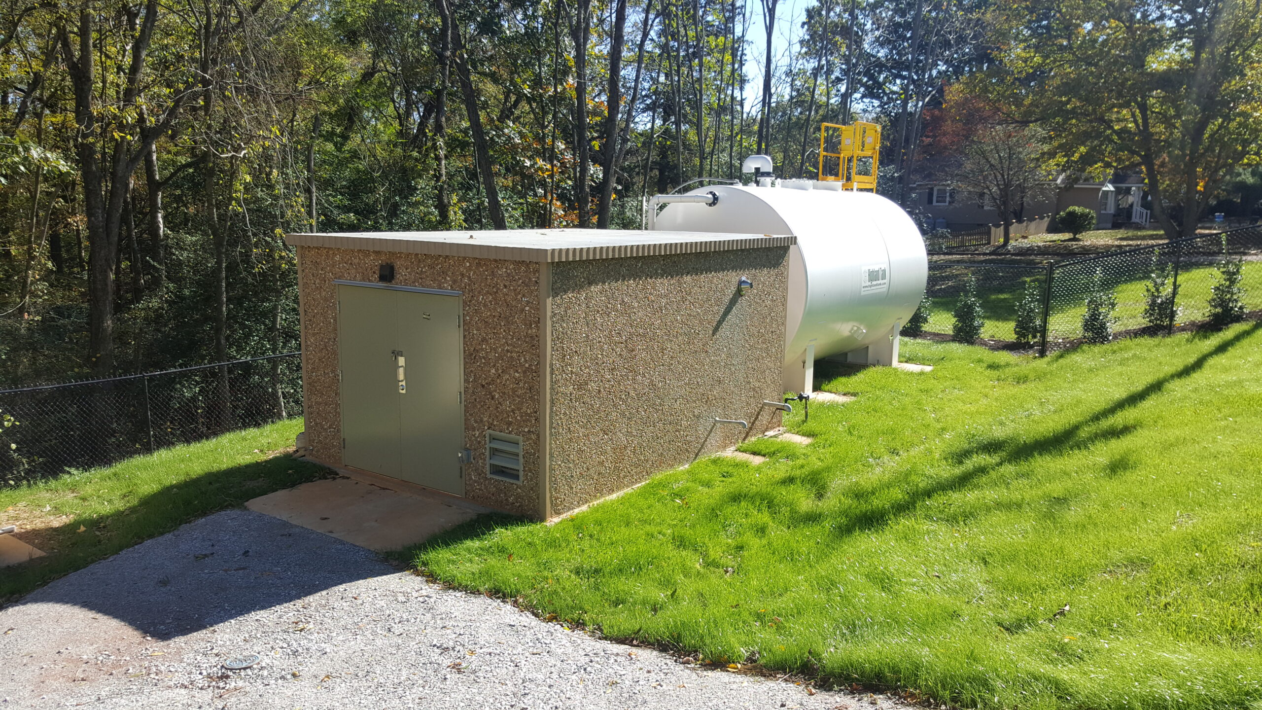 A white cylindrical fuel tank and a brick building are set on well-maintained green grass, enclosed by a chain-link fence amidst tall trees under a bright sky.
