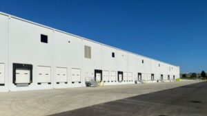 A white warehouse building with black windows, set against a clear sky. The surrounding area is empty, featuring concrete ground and parking spaces. The structure appears unused and abandoned.