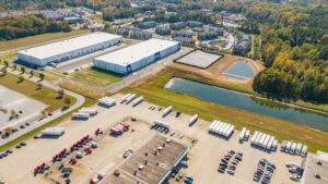 Aerial view of a large parking lot surrounded by water and buildings, featuring vehicles parked in organized rows, with green trees and urban infrastructure visible nearby.