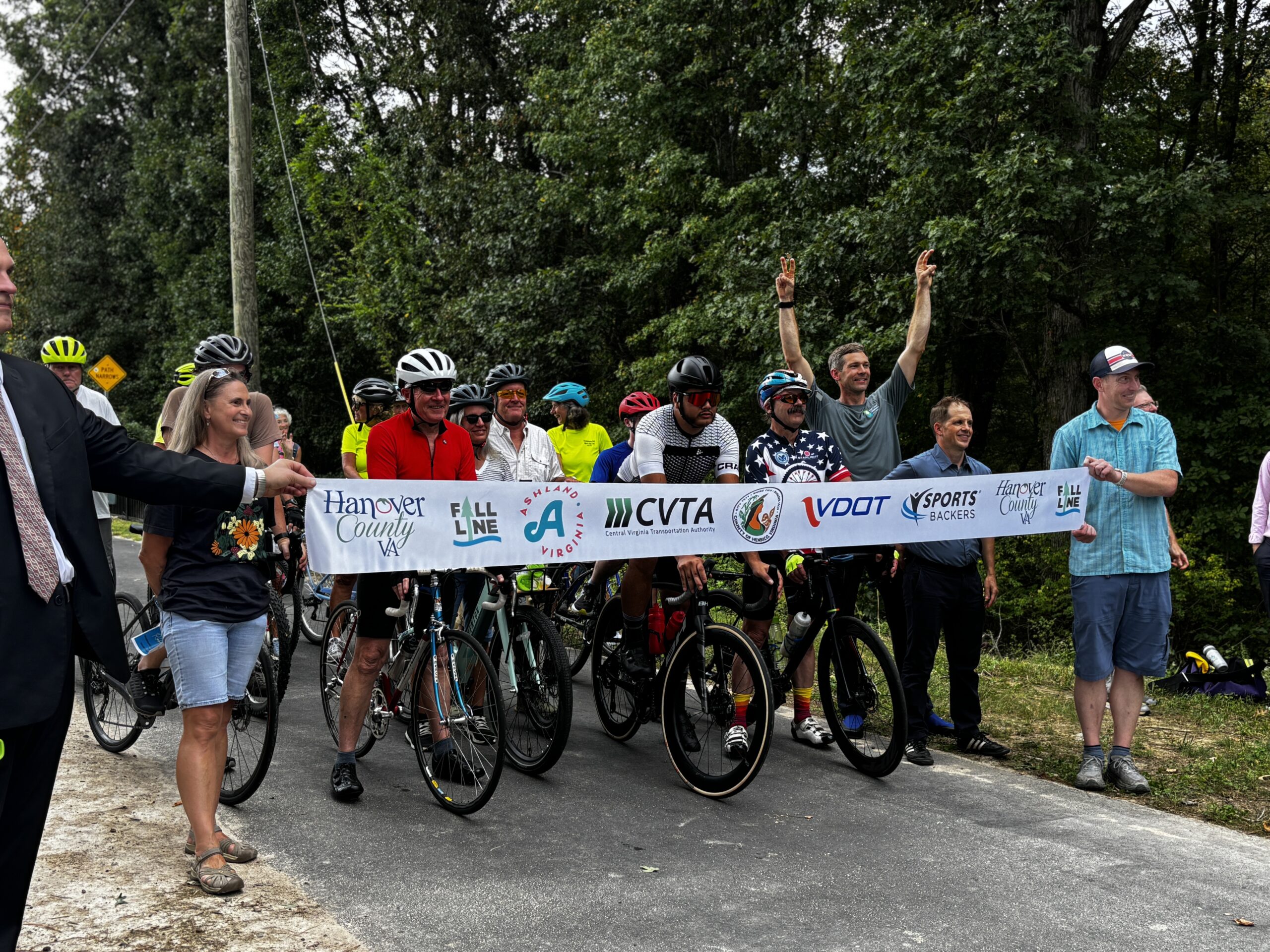 An excited group of people on bicycles waiting behind a banner lined with logos including Hanover County, Virginia, Fall Line, Ashland, Virginia, CVTA, VDOT, and Sports Backers.