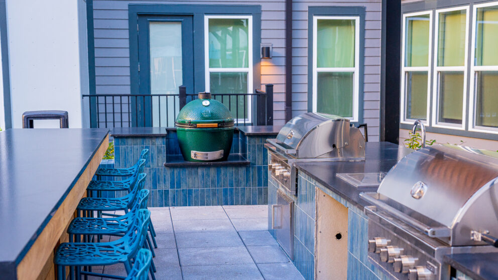 A blue tile patio area with a bar and blue chairs lined up beside it. There are two stainless steel grills and a sink to the bar's right.