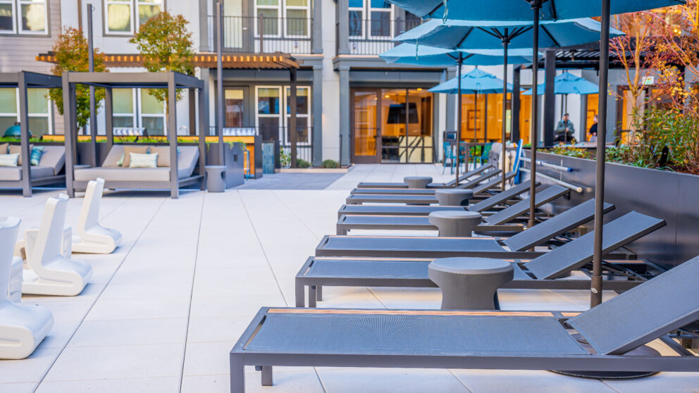 A row of lawn chairs with umbrellas next to them. An apartment building is in the background.