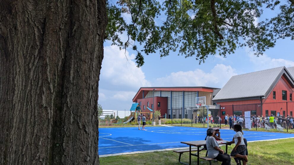 People standing on a basketball court with people sitting at a picnic table next to it.